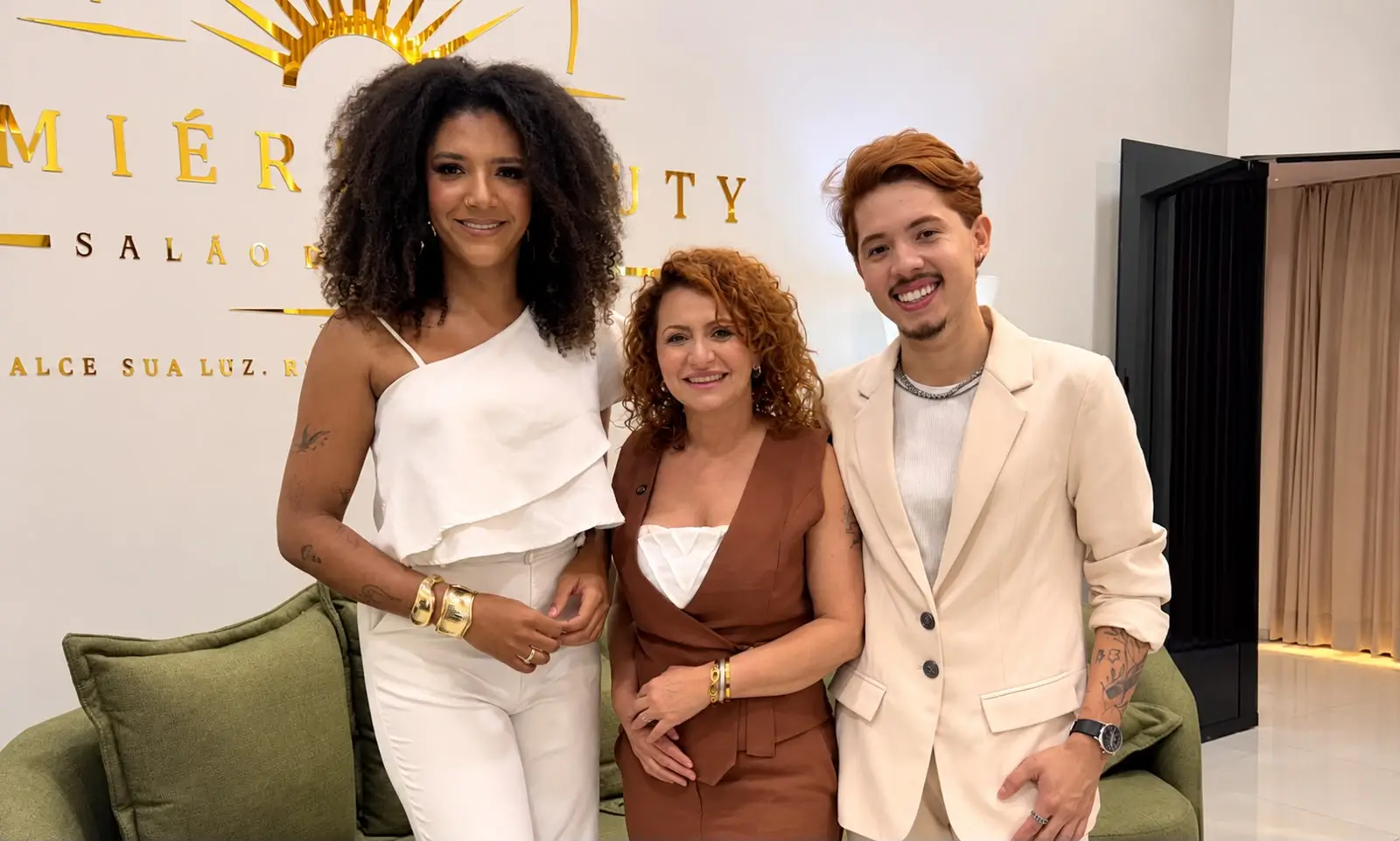 Fotografia em plano médio de três pessoas sorridentes no interior do salão Lumière Beauty. À esquerda, uma mulher de cabelo escuro e encaracolado veste um conjunto branco; ao centro, uma mulher de cabelo ruivo usa um colete castanho; e à direita, um homem de fato bege. Ao fundo, destaca-se a parede branca com o logótipo em letras douradas "LUMIÈRE BEAUTY" sob um emblema de sol estilizado.