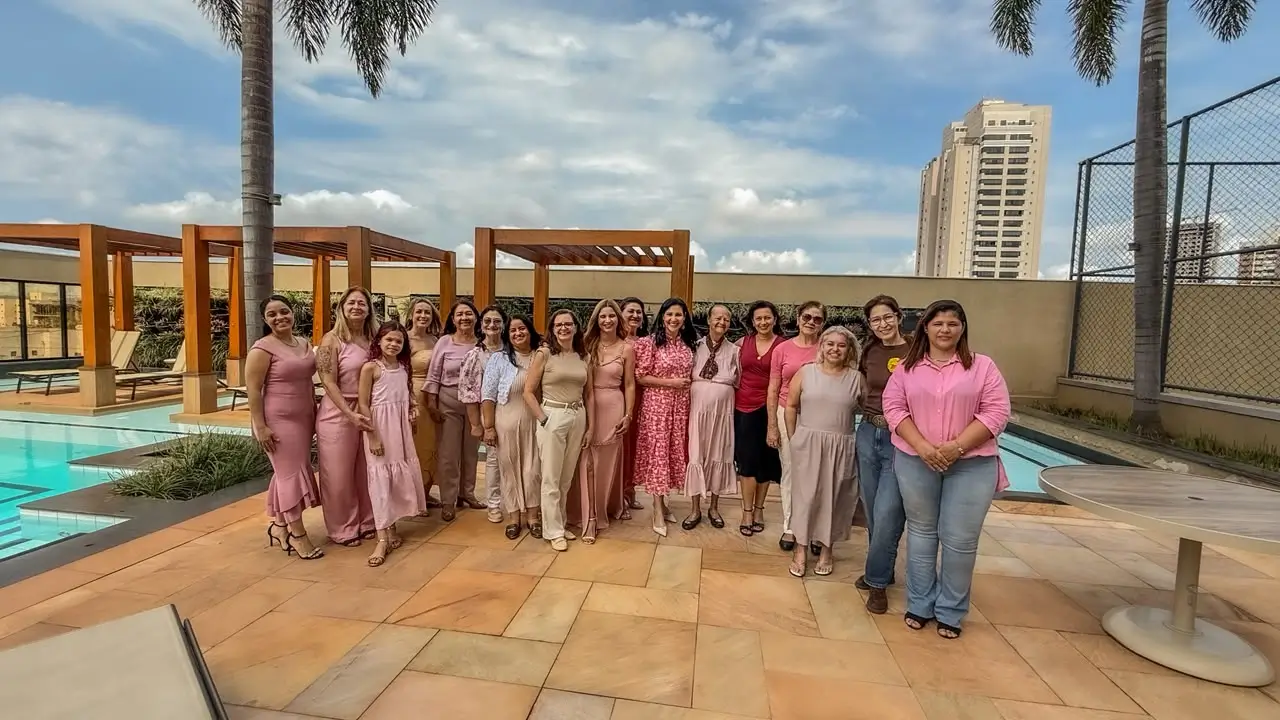 Fotografia aberta ao ar livre, em um terraço com piso de cerâmica e piscina, mostrando um grupo de cerca de vinte mulheres de diversas idades reunidas em pé para uma foto comemorativa. A maioria veste roupas nas cores rosa e tons de nude. Ao fundo, destacam-se estruturas de madeira, palmeiras, uma piscina com água azul e edifícios altos sob um céu parcialmente nublado.