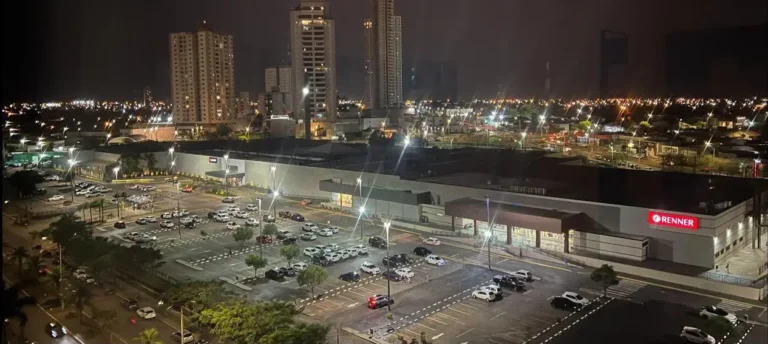 Fotografia aérea noturna do Rondon Plaza Shopping, em Rondonópolis. A imagem mostra a fachada e a ampla área externa do centro de compras iluminadas à noite, destacando o grande estacionamento ao ar livre preenchido por veículos e a entrada principal com o letreiro luminoso da loja Renner em destaque na cor vermelha. Ao fundo, edifícios residenciais altos e iluminados compõem a paisagem urbana sob o céu escuro.
