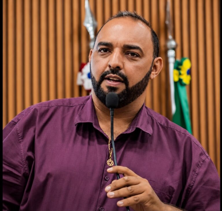 Fotografia em enquadramento fechado do vereador Beto do Amendoim no púlpito da Câmara Municipal. Ele é um homem pardo, de barba e cabelo curto escuros, vestindo uma camisa social de manga comprida na cor roxa e um cordão dourado com pingente de Estrela de David. O parlamentar segura o pedestal do microfone e discursa direcionado à frente. Ao fundo, observa-se um painel ripado de madeira e detalhes de bandeiras oficiais.
