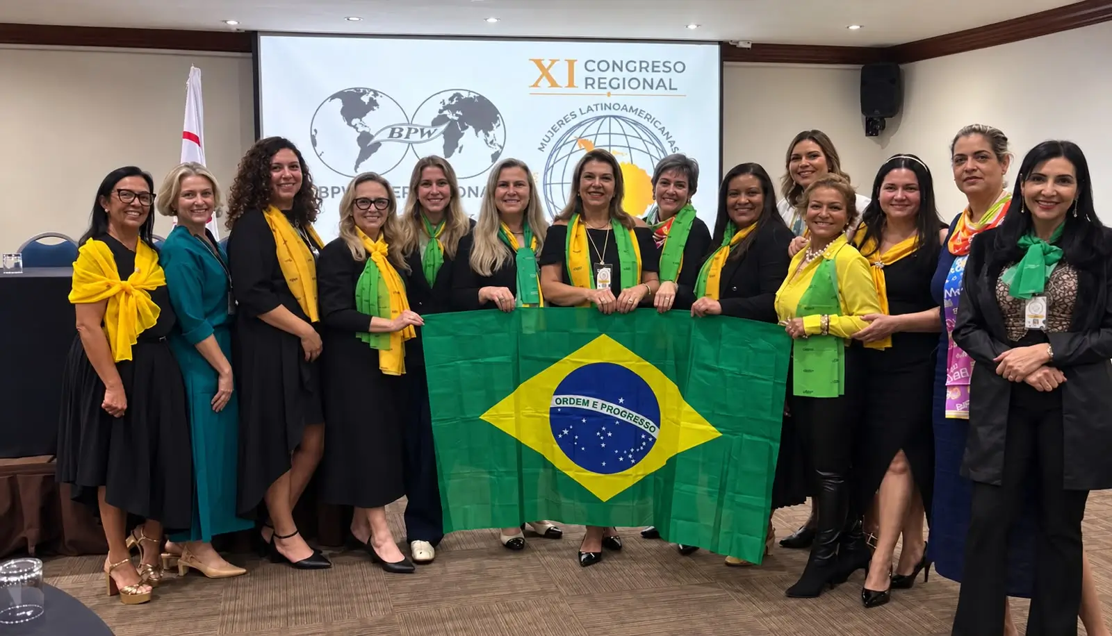 Fotografia interna de um grupo com cerca de quinze mulheres reunidas em pé, sorrindo e posando para a câmera. Ao centro, duas mulheres seguram abertamente a bandeira do Brasil. Várias participantes vestem echarpes nas cores verde e amarela. Ao fundo, vê-se uma tela projetada com o logotipo do evento "XI Congreso Regional - Mujeres Latinoamericanas" e o emblema da BPW, em um auditório ou sala de reuniões.