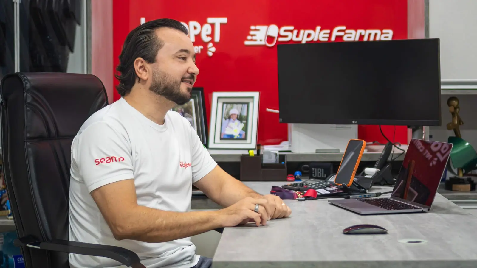 Fotografia em plano médio de Rodolfo Jaquinta sentado numa mesa de escritório cinza. Homem de cabelo escuro, barba e bigode, veste uma t-shirt branca de mangas curtas com logótipos gravados no peito e na manga. Está posicionado de perfil à esquerda, sorrindo e com as mãos apoiadas sobre a mesa. Sobre a banca, veem-se um computador portátil aberto, um telemóvel num suporte, um rato e um monitor. Ao fundo, há uma parede vermelha com marcas impressas como "Suple Farma" e quadros decorativos.