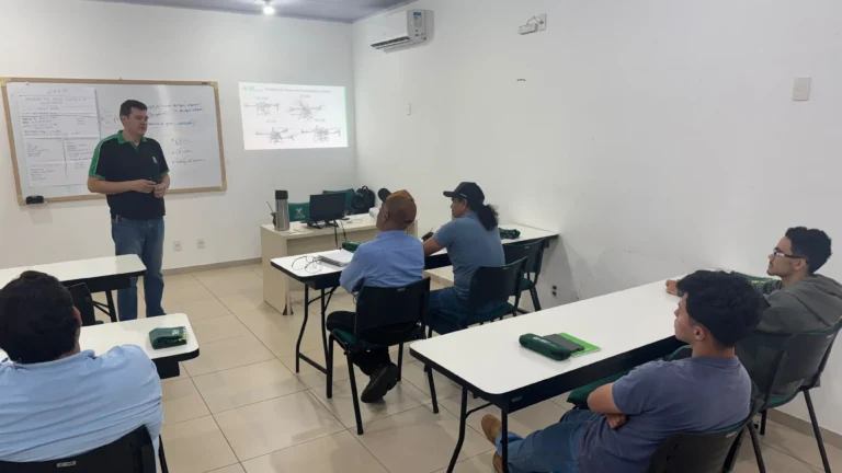 Sala de aula iluminada com paredes brancas e piso claro. À esquerda, em pé, um instrutor vestindo camisa preta com detalhes verdes e calça jeans fala aos alunos. Ao fundo, uma projeção na parede exibe modelos de drones de pulverização agrícola. Vários alunos estão assentados em carteiras brancas dispostas na sala, atentos à explicação.