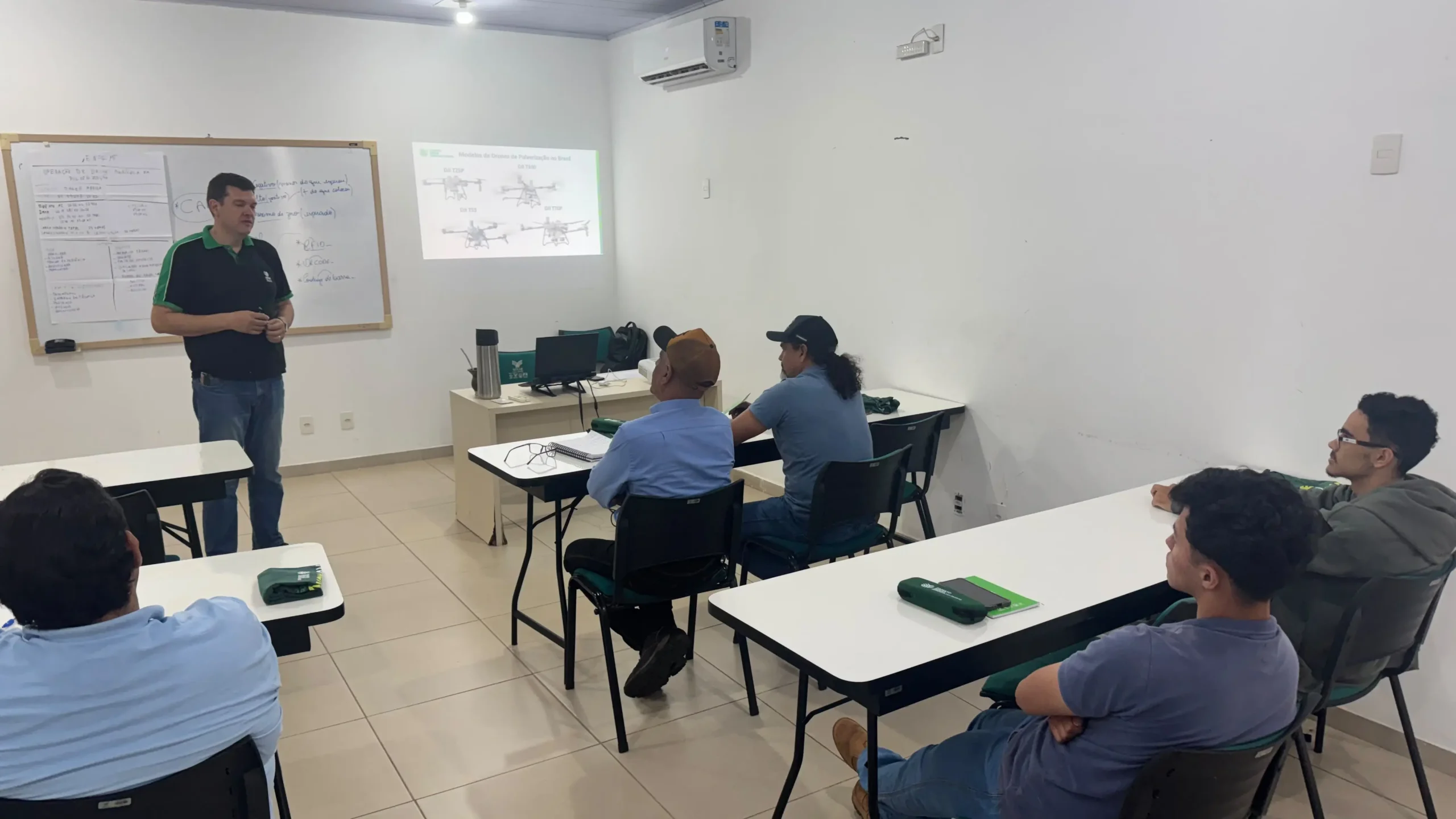 Sala de aula iluminada com paredes brancas e piso claro. À esquerda, em pé, um instrutor vestindo camisa preta com detalhes verdes e calça jeans fala aos alunos. Ao fundo, uma projeção na parede exibe modelos de drones de pulverização agrícola. Vários alunos estão assentados em carteiras brancas dispostas na sala, atentos à explicação.