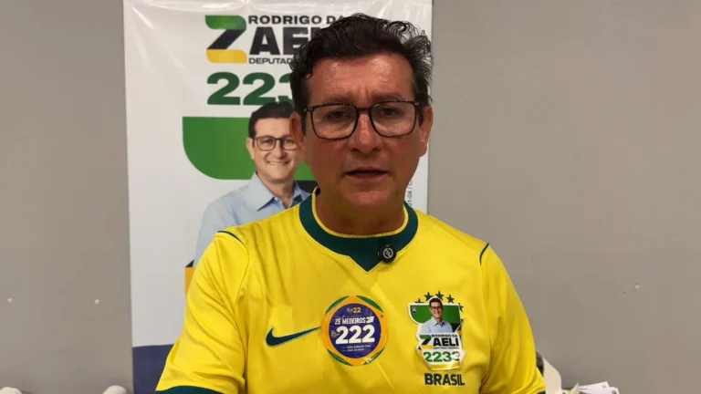 Homem de óculos de armação escura e camisola amarela da seleção brasileira com autocolantes de campanha política, incluindo o seu nome Rodrigo da Zaeli e o número 2233. Ele está posicionado em frente a um painel publicitário com a sua foto e nome de campanha.