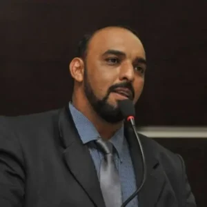 Fotografia em plano fechado do vereador, um homem de pele morena, barba e cabelo curtos escuros, falando ao microfone. Ele veste terno escuro, camisa social azul-escura listrada e gravata cinza.