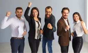 Fotografia em plano inteiro de um grupo de cinco profissionais, três homens e duas mulheres, em trajes sociais e esporte fino, posicionados lado a lado contra um fundo cinza claro. Todos sorriem entusiasmadamente e erguem os punhos em sinal de comemoração e vitória. O primeiro homem à esquerda veste camisa branca e segura papéis; a mulher ao centro veste blazer preto sobre blusa branca; ao lado dela, um homem de barba e óculos veste blazer escuro e calça de ganga; o quarto homem veste blazer castanho; e a mulher à direita veste camisa branca, calça preta e segura uma agenda.