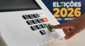 A imagem mostra uma mão humana com o dedo indicador prestes a apertar o botão verde 'CONFIRMA' em uma urna eletrônica. A urna tem um teclado numérico com braille e botões coloridos para 'BRANCO', 'CORRIGE' e 'CONFIRMA'. Ao fundo, à direita, um painel azul exibe a frase 'ELEIÇÕES 2026' e a logo da Justiça Eleitoral, com a frase 'VOTO DEMOCRÁTICO' logo abaixo. A imagem é uma foto em primeiro plano, com foco na urna e na mão do eleitor. O fundo é escuro e a iluminação é suave. Na parte inferior, há ícones de acessibilidade e de um tablet.