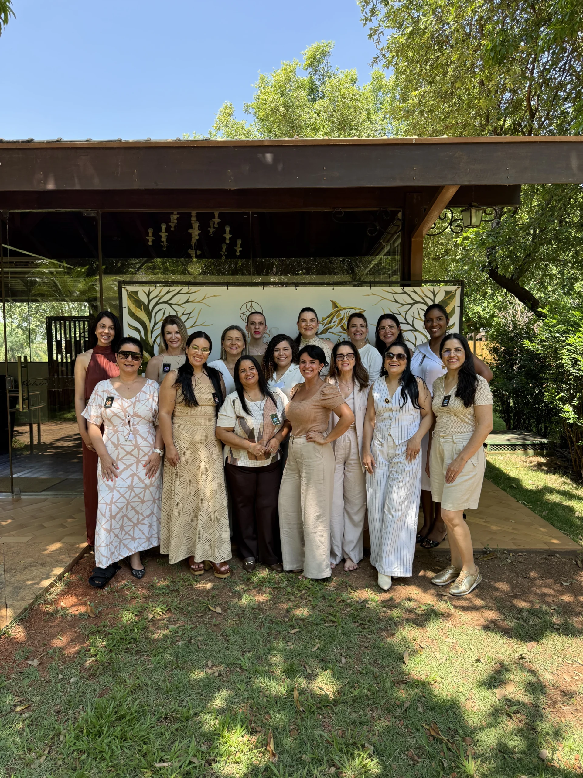 Uma fotografia em plano médio, tirada ao ar livre durante o dia, mostrando um grupo de mulheres sorridentes posando para a foto em grupo. Elas estão organizadas em duas fileiras, na frente de uma estrutura de madeira rústica e vidro que parece um restaurante ou espaço de eventos. O chão é uma mistura de pedra e grama. As mulheres estão vestidas em roupas casuais e elegantes, predominantemente em tons claros e neutros, com algumas em cores mais vibrantes. Muitas usam óculos de sol e crachás de identificação. No fundo, há uma faixa ou banner branco e dourado com um logotipo central e o texto "IMERSÃO LÍDER ÁUREA" e "26.set.2026 - Rondonópolis-MT" visível em dourado. A luz solar é brilhante, criando sombras nítidas. A composição é centralizada e transmite uma atmosfera de união e celebração.
